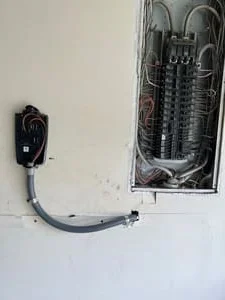 Wall-mounted EV charger on white wall with gray flexible conduit leading to an open electrical panel showing breakers and wiring
