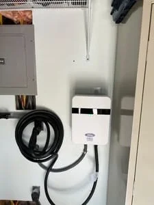 White wall-mounted EV charger with black charging cable beside a gray electrical panel in a garage