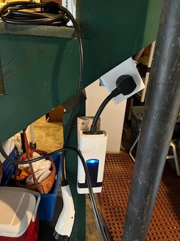 White wall-mounted EV charger with blue indicator light and black charging cable on green wall, beside a dedicated outlet on a white junction box