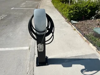 White EV charger on dark metal pedestal with coiled black cable, mounted on concrete beside a parking space