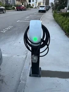 White pedestal EV charger with coiled black cable and green status light mounted on concrete sidewalk beside a street