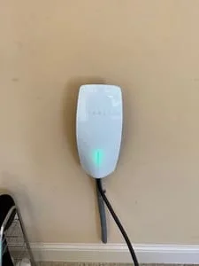 White wall-mounted EV charger with green status light and black charging cable on beige painted wall