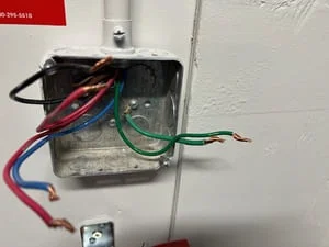 Metal junction box on white wall with black, red, blue, green, and bare copper electrical wires terminated inside, conduit entering from above