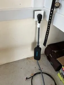 Wall-mounted dark gray EV charger with black cable installed in a garage above a white outlet box