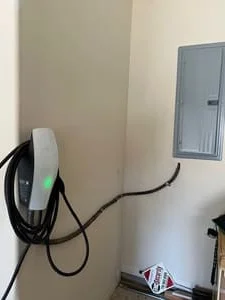 Wall-mounted white EV charger with green indicator light and coiled cable beside a gray electrical panel