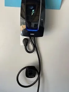 Black wall-mounted EV charger with blue status light and coiled charging cable on white wall
