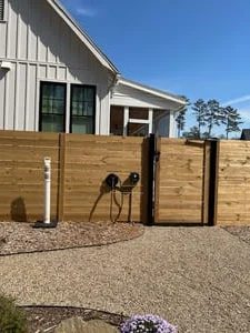 Wall-mounted EV charger with coiled cables beside a horizontal cedar privacy fence and gate, gravel landscaping
