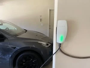 Wall-mounted EV charger with green status light and charging cable connected to a dark gray electric vehicle in a garage
