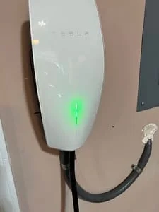 Tesla wall connector EV charger mounted on tan wall next to gray electrical panel with green status light on