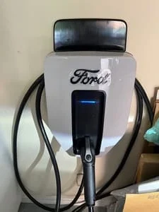 Wall-mounted Ford Level 2 EV charger with white housing, blue status light, and hanging charging cable