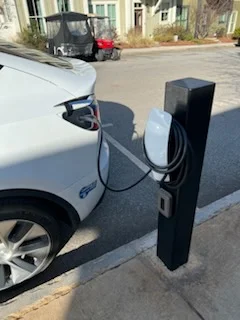 Black pedestal EV charging station with white charging handle and cable connected to a white electric vehicle in a paved parking space