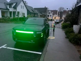 Dark electric pickup truck with green LED light bar parked beside an outdoor EV charging station on a wet residential driveway