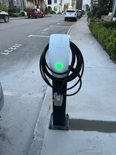 White EV charger with coiled black cable on black pedestal, green status light, concrete sidewalk installation