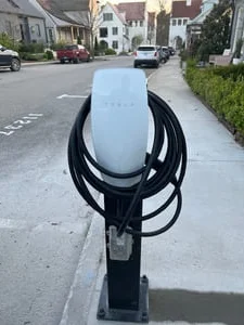 White EV charger with coiled black cable mounted on a black post at a concrete curbside base