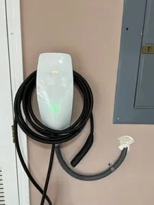 White wall-mounted EV charger with green indicator light and coiled cable beside gray electrical panel
